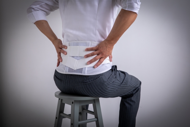 腰に手を添え、サポートベルトを着用した人物の後ろ姿。腰痛を暗示する構図。